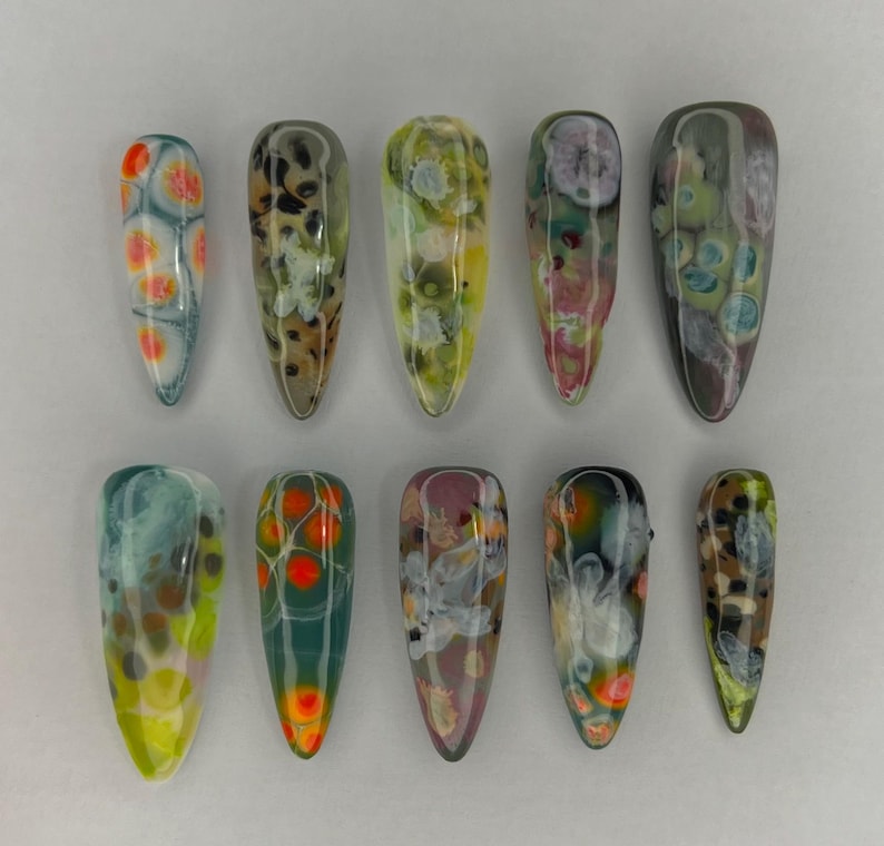 May include: Ten artificial nails with abstract designs. The nails are in various colors, including orange, green, yellow, and brown, with a glossy finish. The shapes are long and pointed, resembling a stiletto style.