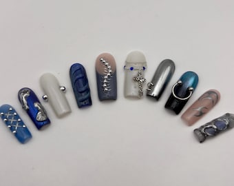 May include: A collection of ten artificial nails with various designs. The nails feature colors like blue, silver, black, and pink, with embellishments such as silver beads, swirls, and a cross pendant. Some nails have a metallic finish, while others have a matte appearance.