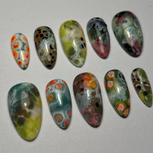 May include: A collection of ten oval-shaped artificial nails. Each nail features a unique abstract design with a mix of colors, including orange, green, brown, and white. The designs resemble a marbled or speckled pattern.