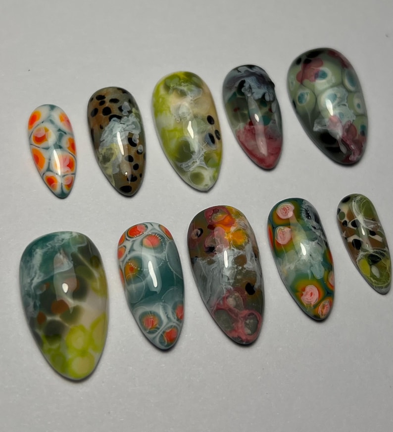 May include: A collection of ten oval-shaped artificial nails. Each nail features a unique abstract design with a mix of colors, including orange, green, brown, and white. The designs resemble a marbled or speckled pattern.