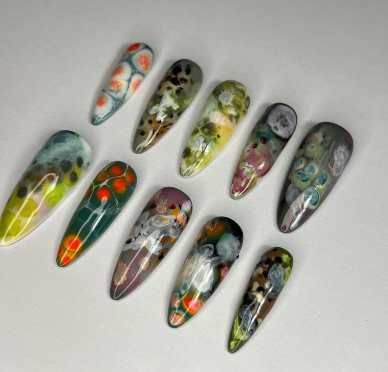 May include: A collection of ten long, almond-shaped artificial nails. Each nail features a unique abstract design with a glossy finish. Colors include green, orange, yellow, blue, and purple, creating a vibrant and artistic display.