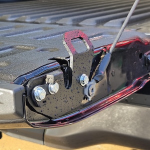 May include: A black metal tailgate latch assembly for a pickup truck, featuring a textured finish with visible water droplets. The component includes a vertical bracket with a rectangular cutout, secured by three silver hexagonal bolts to the truck bed.