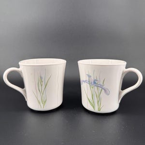 May include: Two white ceramic mugs with curved handles. Each mug features a floral design with green and purple iris flowers. The mugs are set against a dark background, highlighting the delicate floral details.