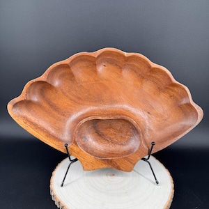 Könnte beinhalten: Ein Holzserviertablett in Fächerform. Das hellbraune Holztablett hat einen gewellten Rand und eine zentrale, ovale Vertiefung. Es wird auf einem schwarzen Metallständer und einer Holzscheibe präsentiert.