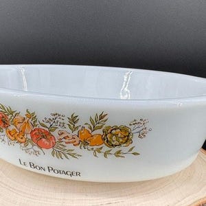 May include: White oval casserole dish with handles, decorated with a colorful vegetable and herb design. The words "LE BON POTAGER" are printed on the front. The dish is sitting on a wooden surface.