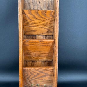 May include: A wooden wall-mounted organizer with three tiered pockets and three small brass hooks. The organizer is made of light brown wood with visible wood grain. The design is rectangular with a rustic aesthetic.