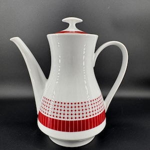 May include: White ceramic coffee pot with a red geometric pattern around the base and on the lid. The pot has a long spout and a curved handle. The design is a retro style, perfect for serving coffee or tea.