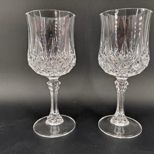 May include: Two clear crystal wine glasses with intricate diamond and vertical line patterns. The glasses have a tulip-shaped bowl, a slender stem, and a round base. The background is a dark gray.