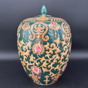 May include: A dark green ceramic jar with a matching lid featuring a pointed finial. The surface is decorated with intricate, raised golden-brown scrollwork and floral patterns, accented with small, vibrant pink teardrop-shaped details.