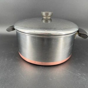 Puede incluir: Una olla redonda con un exterior de metal gris oscuro cepillado y una base de color cobre. La olla cuenta con dos asas laterales negras y una tapa circular a juego con un pomo central negro, presentando un utensilio de cocina funcional.