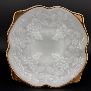 May include: A white, oval-shaped serving dish with a gold rim. The dish features a raised grape and leaf design around the edges. The dish is likely made of glass or porcelain.