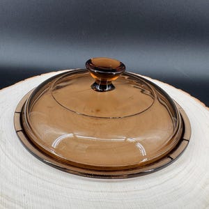 Può includere: Un coperchio di vetro marrone con pomello abbinato, progettato per adattarsi a un piatto rotondo. Il coperchio è trasparente, consentendo la visione del contenuto. Il coperchio è su una superficie di legno chiaro.