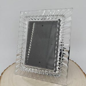 May include: A rectangular picture frame made of clear, transparent glass with a decorative, faceted, and ribbed border design. The frame features a dark gray backing insert and is designed to hold a photograph in a vertical, portrait orientation.