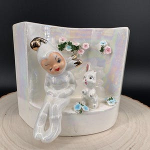 May include: A white ceramic planter with a pearlescent finish, featuring a seated figurine with a gold-tipped hat and a small white rabbit. The planter is decorated with small floral accents in pink and blue.
