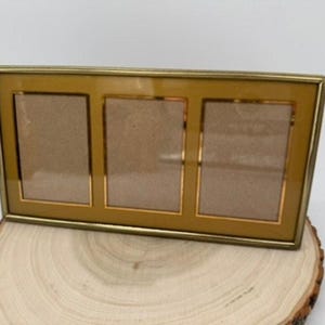 May include: A rectangular triple photo frame with a thin metallic gold finish border. The frame features three vertical openings set against a mustard yellow matting, protected by a clear glass front, designed to hold three separate photographs.
