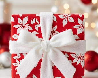 May include: A wrapped gift box with red wrapping paper featuring a white floral pattern. The box is tied with a wide, fluffy white ribbon and bow. The background includes Christmas ornaments and soft, blurred lights.
