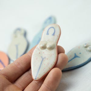 May include: A collection of small, flat, ceramic figurines shaped like stylized human torsos. Each piece features a smooth, glazed finish with speckled details, a raised chest area, and a painted blue line design. The colors include white, peach, and light blue.