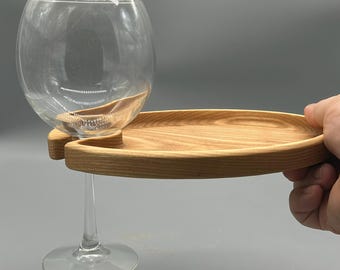 May include: A clear wine glass rests in a wooden wine glass holder. The holder is a light brown color and has a curved design. The glass is empty and has a long stem. The holder is being held by a hand.