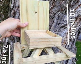 May include: A wooden bird feeder with a picnic table design. The feeder is 10 inches high, 8.25 inches wide, and 7.5 inches deep. It is made of unfinished wood and is designed to be mounted on a tree.