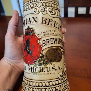 May include: Cream-colored beer stein with a decorative design and the words "IAN BEER BREWING" printed on the side. The stein features a red crest with a black lion and the text "MICH. U.S.A."