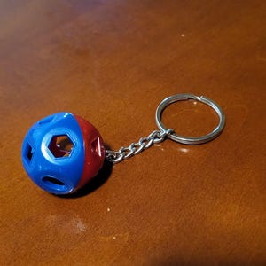 May include: A blue and red ball-shaped keychain with a silver chain and keyring. The ball has hexagonal and circular cutouts. The keychain is on a brown wooden surface.