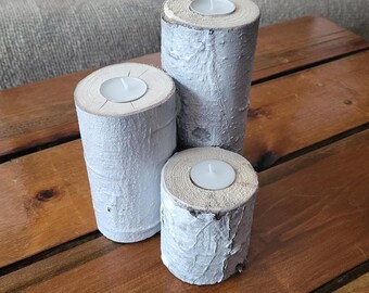 May include: Three white birch wood candle holders with a white tea light candle in each. The candle holders are stacked on top of each other on a wooden surface.
