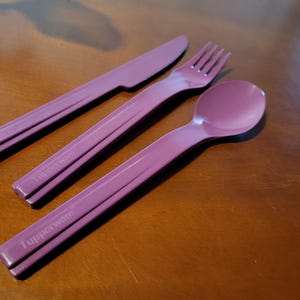 May include: A set of three matching plum-colored Tupperware utensils: a knife, fork, and spoon. The knife and fork have a ridged handle design, and the spoon has a rounded bowl. The Tupperware logo is printed on each handle.