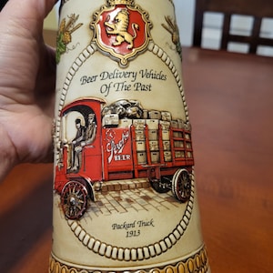 May include: A beige beer stein with a decorative design featuring a red truck labeled "Packard Truck 1913" and the text "Beer Delivery Vehicles Of The Past". The stein has a lion crest and gold accents.