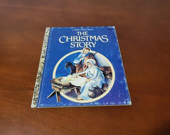 May include: A vintage children's book titled "The Christmas Story" with a blue cover. The cover features an illustration of the Nativity scene within a circular frame. The book's spine has gold accents.