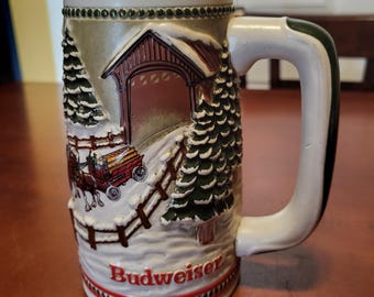 May include: A ceramic beer mug with a winter scene featuring a covered bridge, horse-drawn carriage, and evergreen trees. The mug is white with a detailed relief design and the word "Budweiser" in red.
