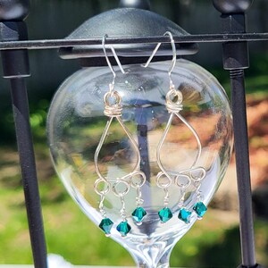 May include: Silver-toned wire earrings with a teardrop design, accented with small, faceted, emerald-green beads. The earrings are displayed against a blurred background, showcasing their delicate craftsmanship and vibrant color.