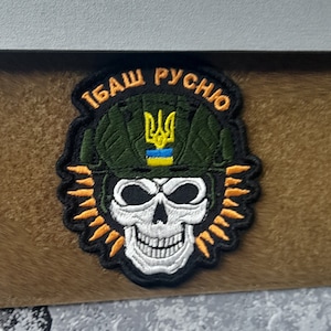 Peut inclure: Écusson brodé représentant un crâne blanc avec des contours noirs, portant un casque vert avec un trident jaune et des rayures bleues et jaunes. Le patch porte l'inscription "TBASH PYCHO" en lettres jaunes.