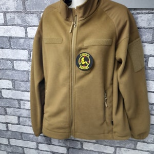 A tan fleece jacket with a zipper closure. The jacket has a patch on the left chest with a black circle and a yellow image of a woman sitting down. The text 'GIRL POWER' is written in black letters on the patch.