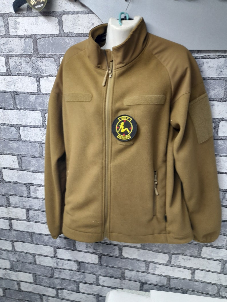 A tan fleece jacket with a zipper closure. The jacket has a patch on the left chest with a black circle and a yellow image of a woman sitting down. The text 'GIRL POWER' is written in black letters on the patch.