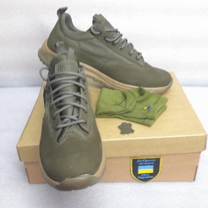 Op de afbeelding: Twee paar olijfgroene suède sneakers met grijze veters in een bruine doos met een Oekraïense vlag en de tekst "help Oekraïne" erop. De doos bevat ook een paar olijfgroene sokken.