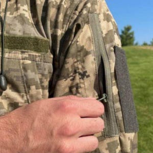 May include: Close-up of a camouflage jacket with a zipper pocket on the sleeve. The jacket has a digital camouflage pattern in shades of green, brown, and tan. A hand is reaching for the zipper pull.