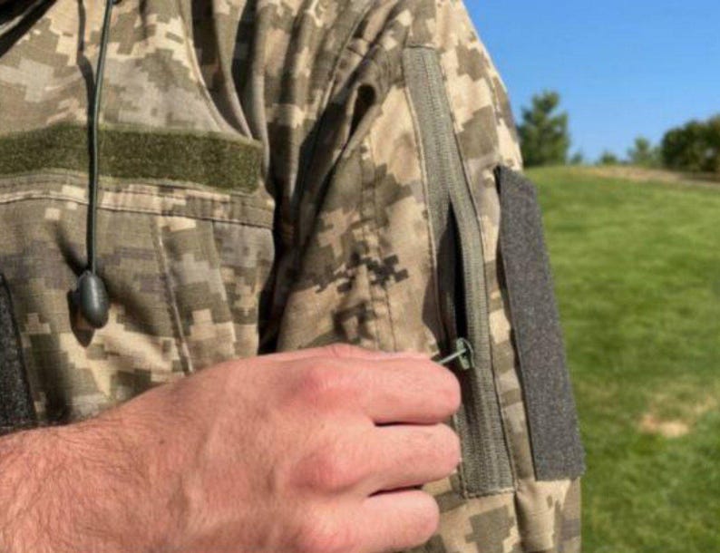 May include: Close-up of a camouflage jacket with a zipper pocket on the sleeve. The jacket has a digital camouflage pattern in shades of green, brown, and tan. A hand is reaching for the zipper pull.