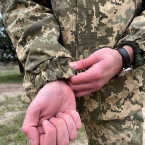 May include: Close-up of a camouflage jacket with a digital pixel pattern in shades of green, brown, and tan. The jacket's sleeve is gathered at the wrist. A black leather watch is visible.