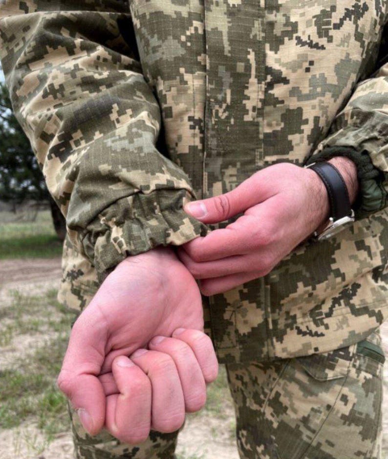 May include: Close-up of a camouflage jacket with a digital pixel pattern in shades of green, brown, and tan. The jacket's sleeve is gathered at the wrist. A black leather watch is visible.