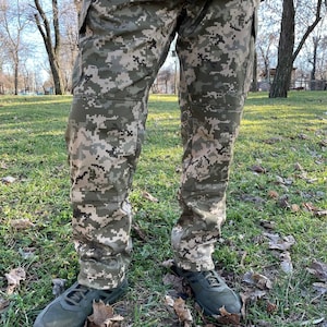May include: A pair of camouflage pants with a digital pattern. The pants have a zipper fly and belt loops. The pants are made of a durable, water-resistant fabric.