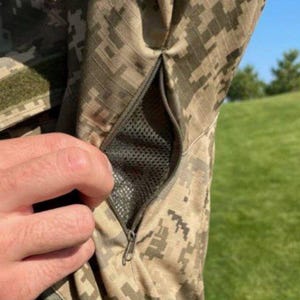 May include: Close-up of a camouflage jacket sleeve with a zippered pocket. The jacket features a digital camouflage pattern in shades of green, brown, and tan. A hand is visible near the pocket, and a green velcro patch is on the sleeve.