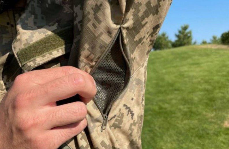 May include: Close-up of a camouflage jacket sleeve with a zippered pocket. The jacket features a digital camouflage pattern in shades of green, brown, and tan. A hand is visible near the pocket, and a green velcro patch is on the sleeve.