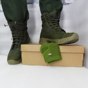Op de afbeelding: Een paar groene militaire laarzen met een bruine kartonnen doos en een groene sok met een gele en blauwe vlag.