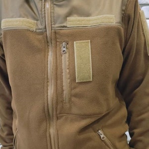 Puede incluir: Una chaqueta de forro polar marr&oacute;n con cierre de cremallera y m&uacute;ltiples bolsillos. La chaqueta tiene un cuello alto y un panel de tela marr&oacute;n contrastante en los hombros.