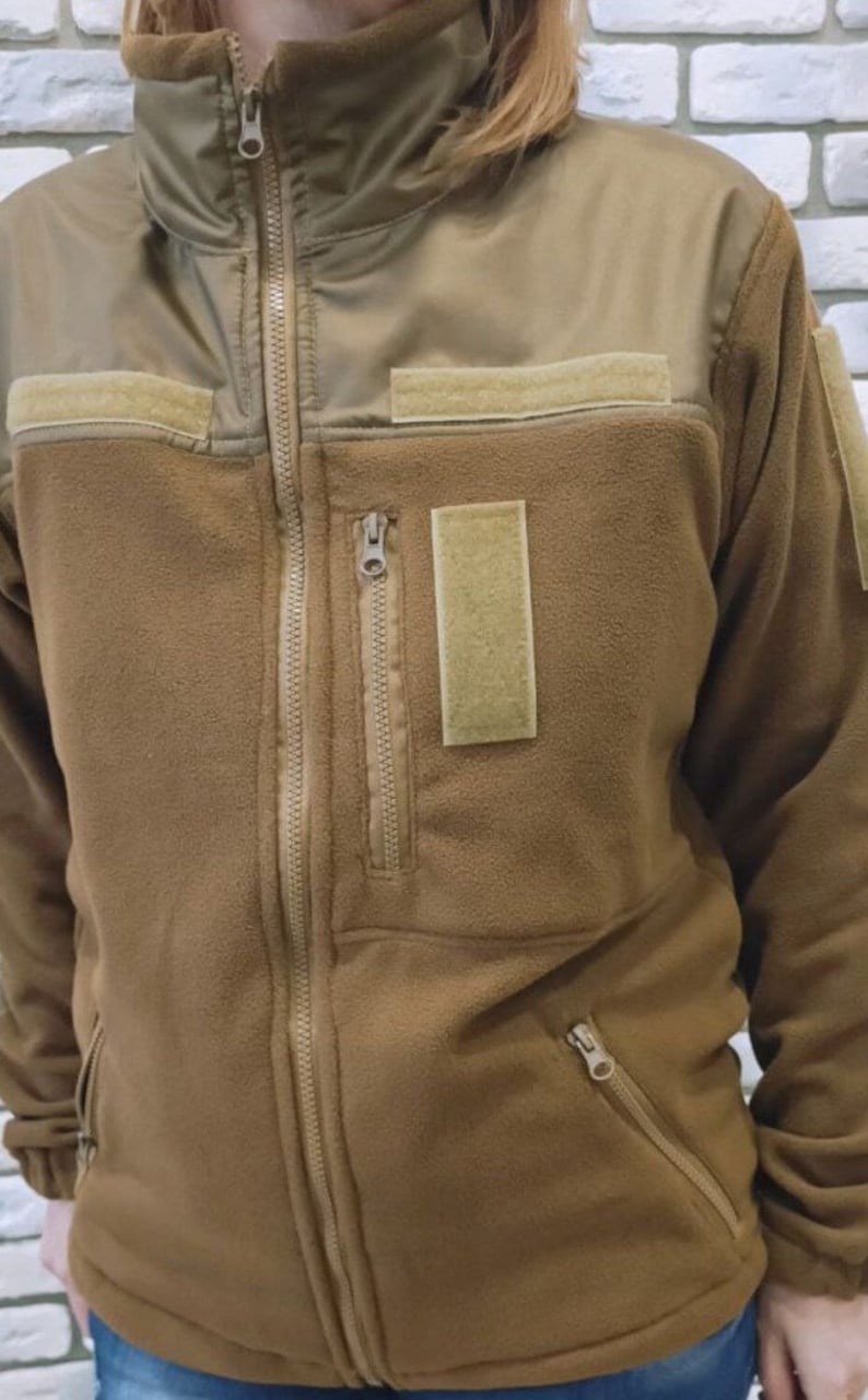 Puede incluir: Una chaqueta de forro polar marr&oacute;n con cierre de cremallera y m&uacute;ltiples bolsillos. La chaqueta tiene un cuello alto y un panel de tela marr&oacute;n contrastante en los hombros.