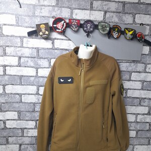 May include: A brown fleece jacket with a zip closure. The jacket has two zipped pockets on the sides and a patch pocket on the left chest. The jacket has a black eye patch on the left chest and a yellow patch on the right sleeve.