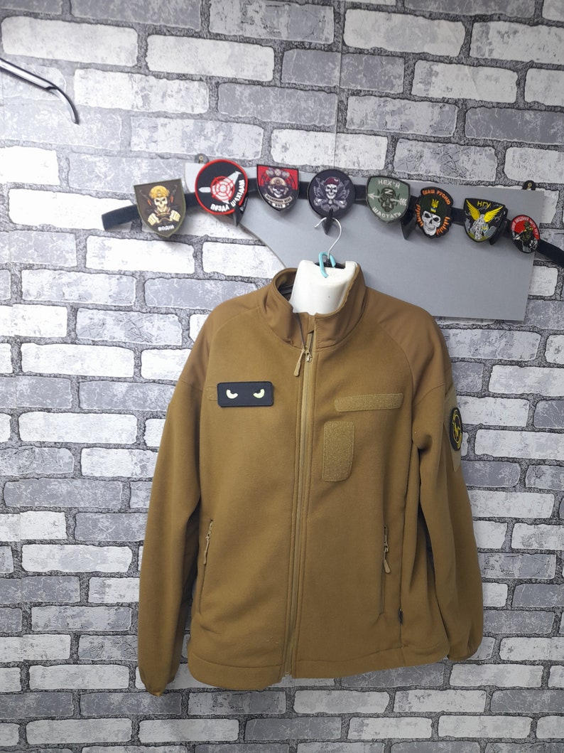 May include: A brown fleece jacket with a zip closure. The jacket has two zipped pockets on the sides and a patch pocket on the left chest. The jacket has a black eye patch on the left chest and a yellow patch on the right sleeve.