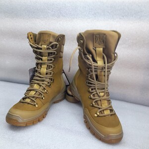 May include: A pair of tan leather hiking boots with a lace-up closure and a black rubber sole. The boots have a yellow logo on the tongue and a yellow strap on the ankle.