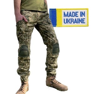 Pode incluir: Uma pessoa vestindo calças camufladas com joelheiras e botas marrons. As calças têm um remendo que diz "Made in Ukraine" com a bandeira da Ucrânia.