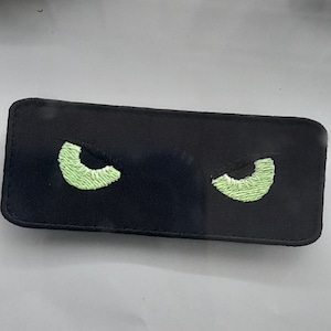 May include: Black fabric patch with two green embroidered eyes. The eyes are shaped like crescent moons.